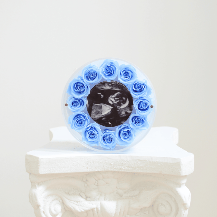 Decorative circular frame with blue preserved roses holding an ultrasound photo on a white pedestal.